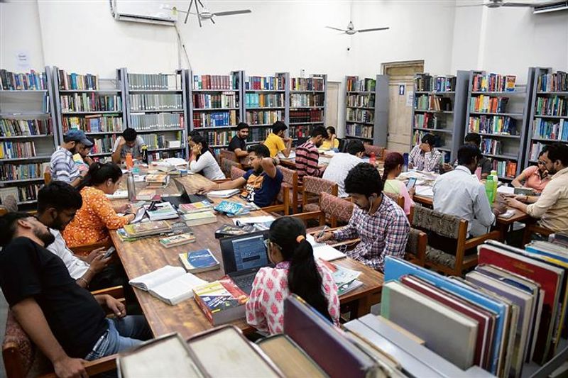 Students utilizing laptops and books in a well-stocked library with extensive bookshelves and wooden study tables.